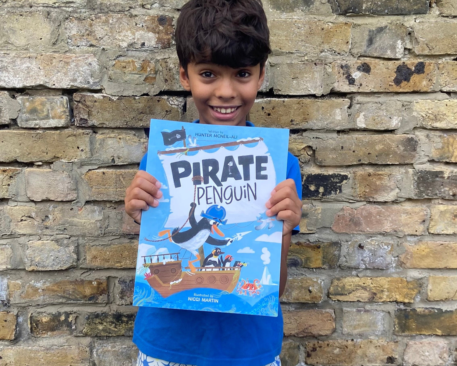 Hunter, a 10-year-old child holding his book Pirate Penguin in front of a brick garden wall